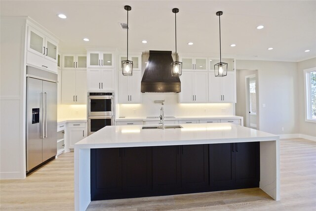 kitchen with white cabinetry, stainless steel appliances, premium range hood, a center island with sink, and light wood-type flooring