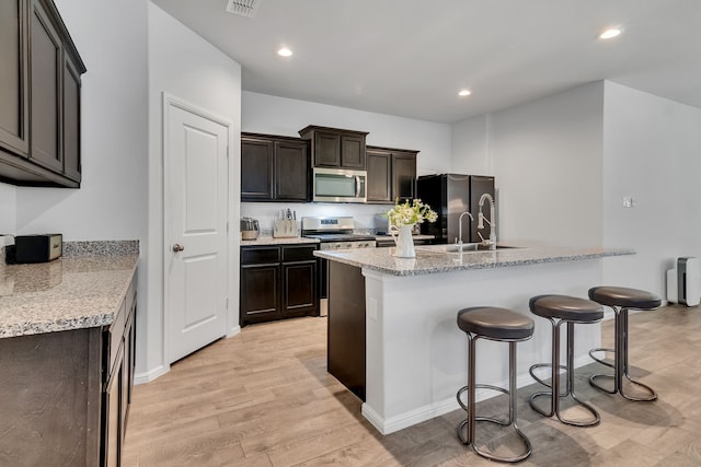 kitchen featuring a kitchen island with sink, dark brown cabinetry, light stone counters, stainless steel appliances, and light hardwood / wood-style flooring