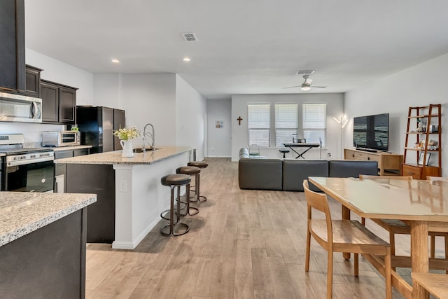 kitchen featuring light stone countertops, dark brown cabinetry, stainless steel appliances, ceiling fan, and a kitchen island with sink