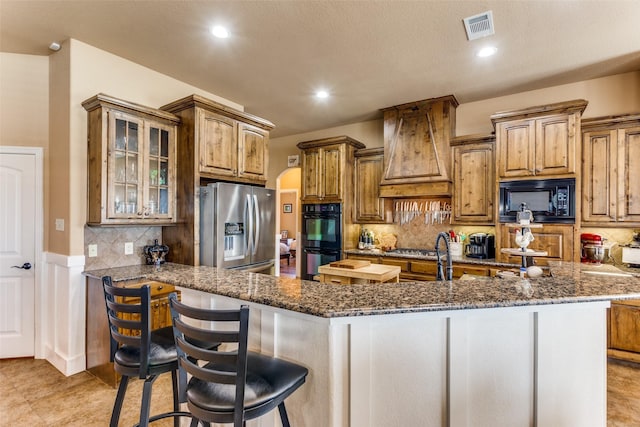 kitchen featuring black appliances, dark stone counters, premium range hood, and a center island with sink