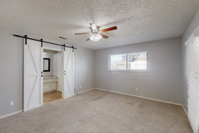 unfurnished bedroom with ensuite bathroom, a textured ceiling, light colored carpet, ceiling fan, and a barn door