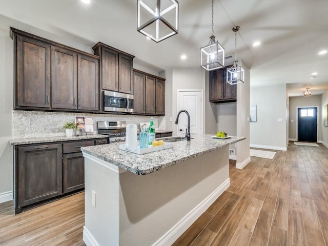 kitchen with an island with sink, sink, hanging light fixtures, light stone counters, and stainless steel appliances