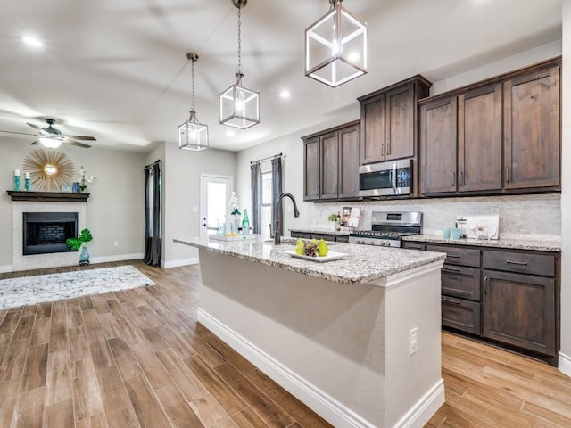 kitchen with light stone counters, dark brown cabinets, a center island with sink, appliances with stainless steel finishes, and pendant lighting