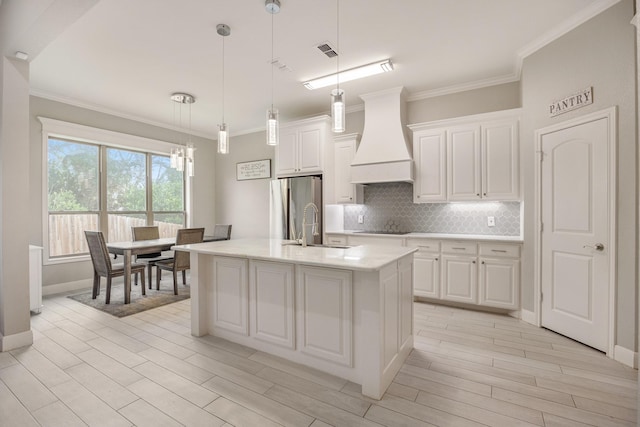 kitchen with premium range hood, stainless steel refrigerator, a center island with sink, and white cabinets