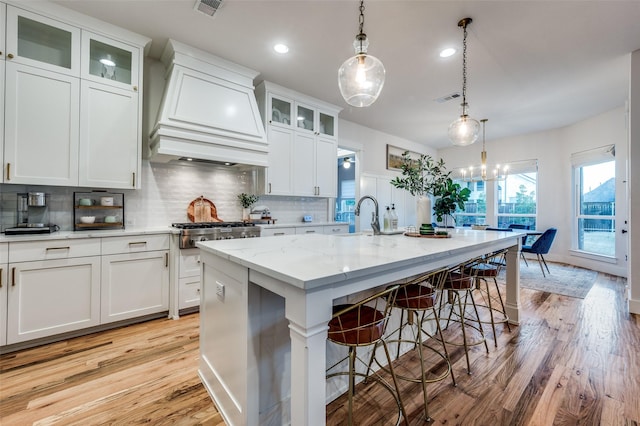 kitchen featuring tasteful backsplash, custom range hood, light hardwood / wood-style flooring, white cabinetry, and an island with sink