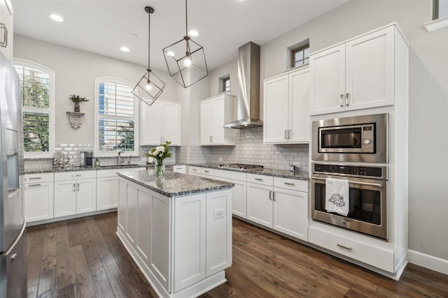 kitchen with wall chimney exhaust hood, stainless steel appliances, a kitchen island, dark stone counters, and white cabinets