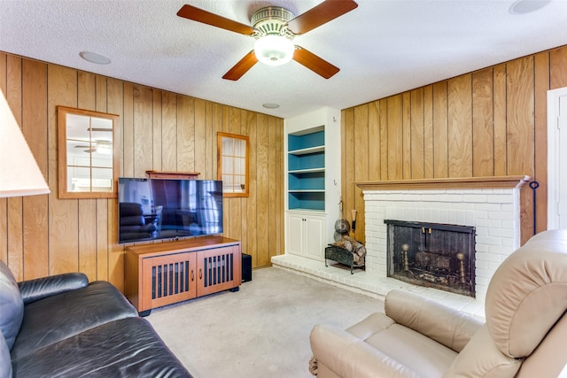 carpeted living room featuring a brick fireplace, built in shelves, a textured ceiling, ceiling fan, and wooden walls