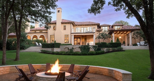 back house at dusk with a lawn, a balcony, and an outdoor fire pit