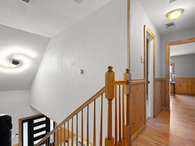 hall with light wood-style flooring, wooden walls, an upstairs landing, visible vents, and wainscoting