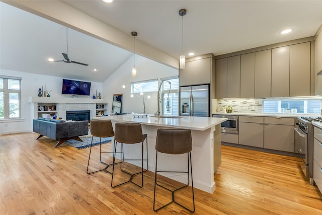 kitchen featuring gray cabinetry, ceiling fan, stainless steel appliances, a brick fireplace, and a kitchen island with sink