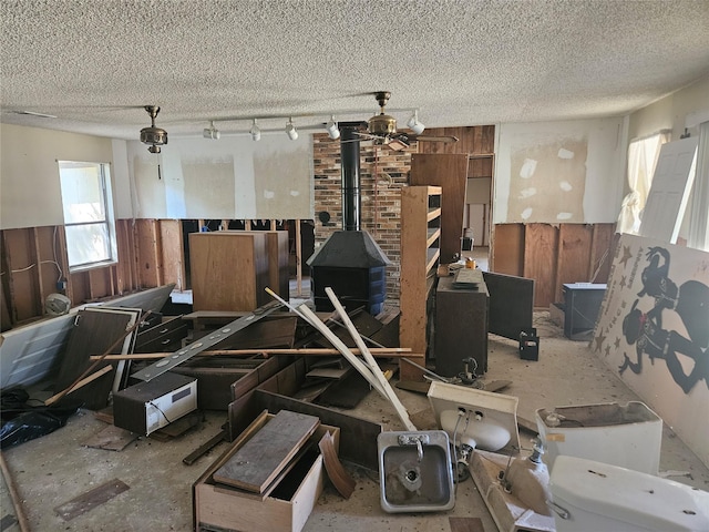 miscellaneous room featuring wood walls, a wood stove, rail lighting, ceiling fan, and a textured ceiling