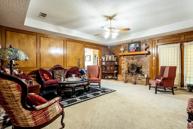 living room with a brick fireplace, ceiling fan, a textured ceiling, a tray ceiling, and light colored carpet