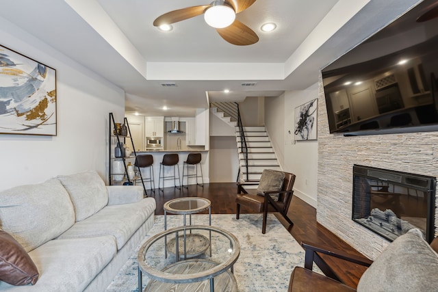 living room with a raised ceiling, ceiling fan, a fireplace, and dark wood-type flooring