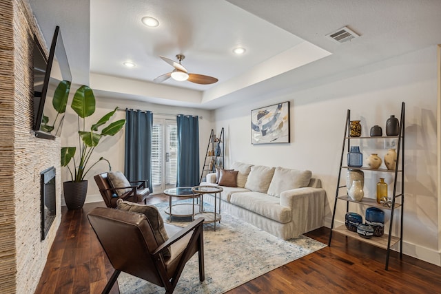 living room featuring ceiling fan, a stone fireplace, a raised ceiling, and dark hardwood / wood-style flooring