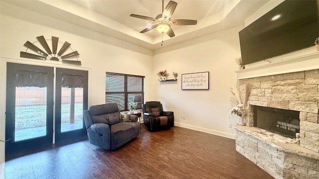 living area with a raised ceiling, a stone fireplace, dark wood-type flooring, and ceiling fan