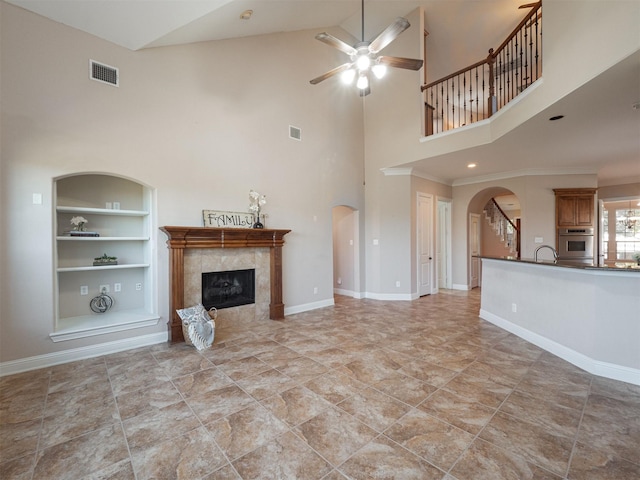 unfurnished living room with a high ceiling, sink, built in shelves, ceiling fan, and a tiled fireplace