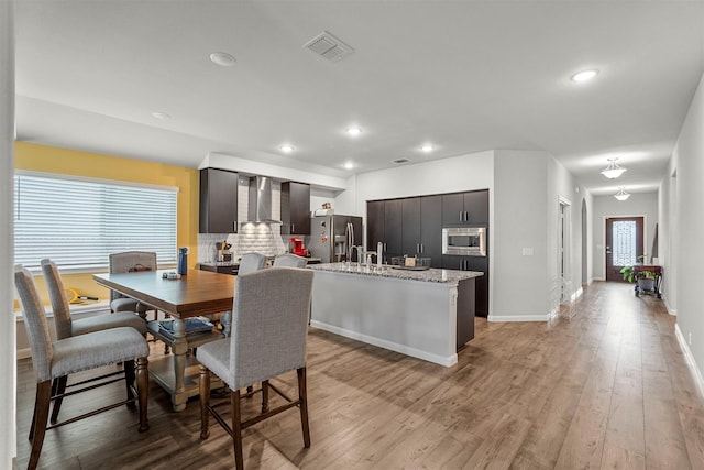 kitchen with wall chimney range hood, stainless steel appliances, light stone countertops, a center island with sink, and light wood-type flooring