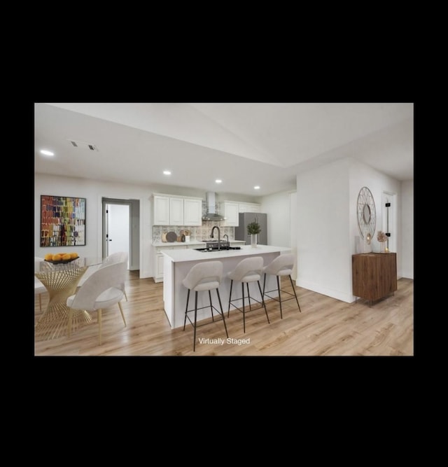 kitchen with wall chimney exhaust hood, stainless steel fridge, a breakfast bar area, a center island with sink, and white cabinets