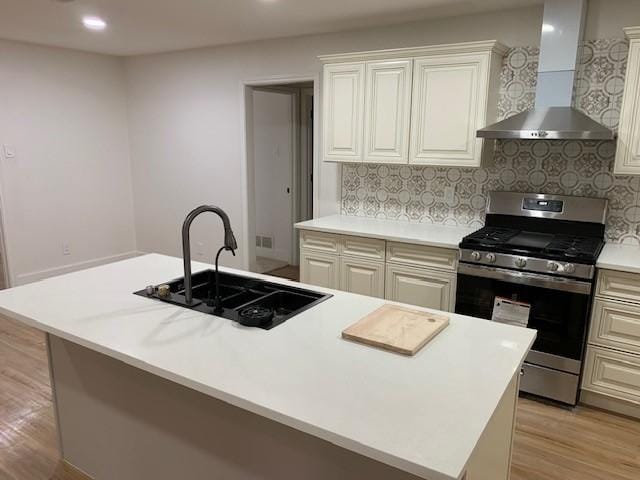 kitchen with sink, stainless steel range with gas stovetop, an island with sink, wall chimney exhaust hood, and light wood-type flooring