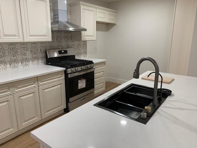 kitchen with sink, wall chimney range hood, stainless steel gas range, decorative backsplash, and white cabinets