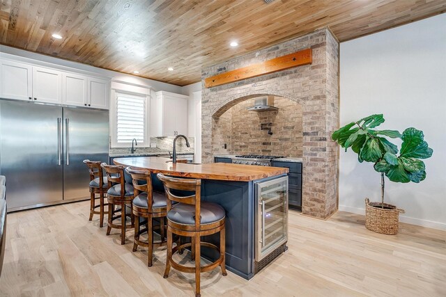 kitchen featuring wood counters, a kitchen island with sink, built in refrigerator, white cabinetry, and wood ceiling