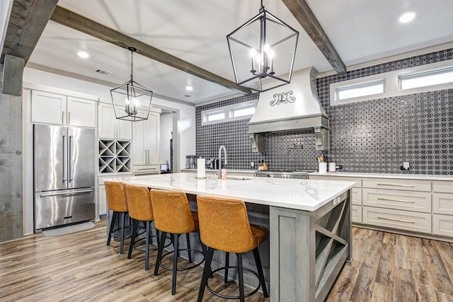 kitchen featuring appliances with stainless steel finishes, custom range hood, sink, hanging light fixtures, and an island with sink