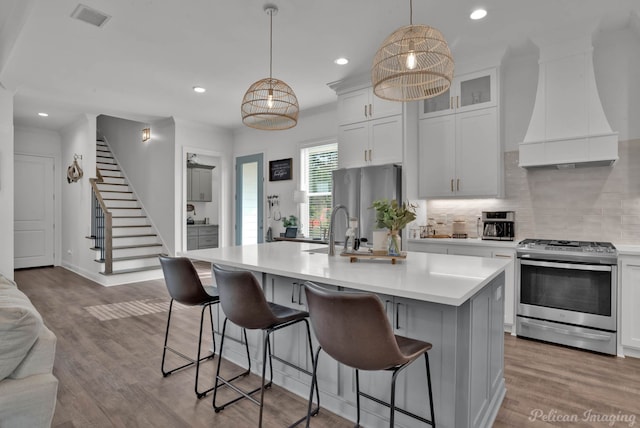 kitchen featuring appliances with stainless steel finishes, decorative light fixtures, white cabinetry, an island with sink, and custom range hood