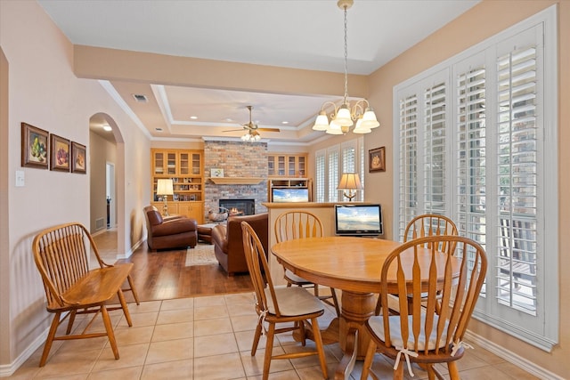 tiled dining space featuring built in shelves, a brick fireplace, a tray ceiling, ceiling fan with notable chandelier, and ornamental molding