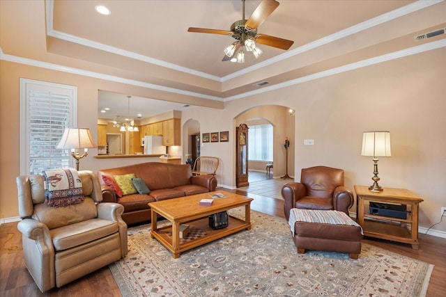 living room with wood-type flooring, ceiling fan with notable chandelier, a raised ceiling, and ornamental molding