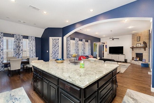 kitchen with dark hardwood / wood-style flooring, light stone counters, ceiling fan, a kitchen island, and a stone fireplace