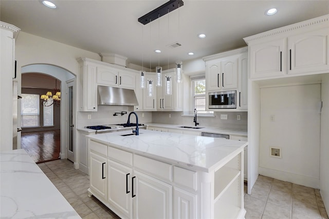 kitchen with appliances with stainless steel finishes, white cabinets, hanging light fixtures, light stone counters, and a center island with sink