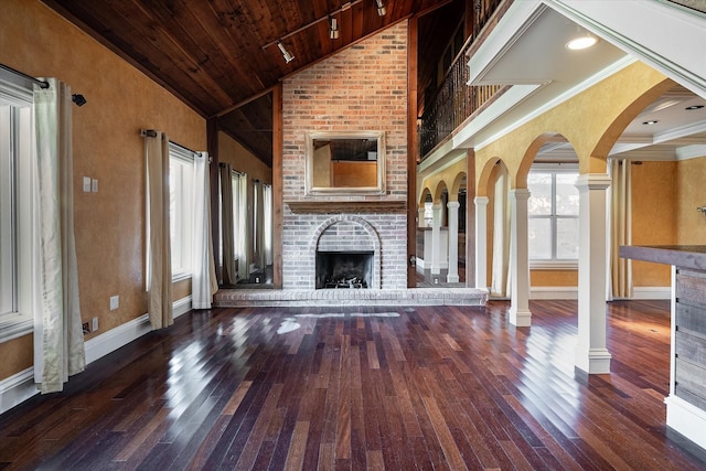 unfurnished living room featuring a brick fireplace, ornate columns, dark wood-type flooring, high vaulted ceiling, and wooden ceiling