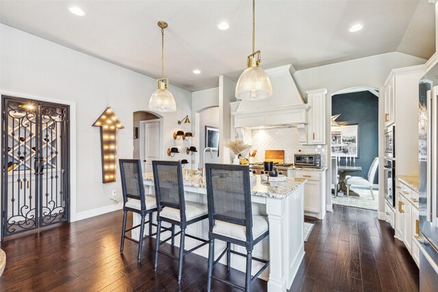 kitchen featuring backsplash, premium range hood, hanging light fixtures, an island with sink, and white cabinetry