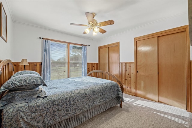 carpeted bedroom with a wainscoted wall, two closets, ornamental molding, a ceiling fan, and wood walls