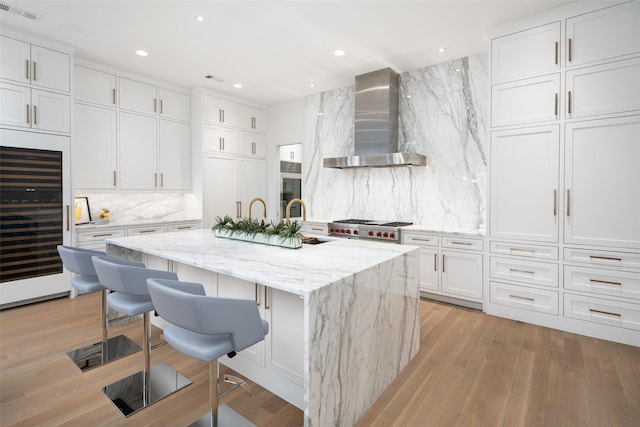 kitchen featuring a breakfast bar, a kitchen island with sink, wall chimney range hood, white cabinetry, and beverage cooler