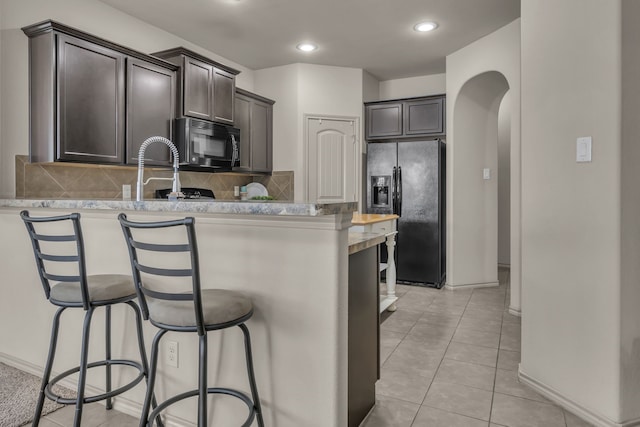 kitchen with tasteful backsplash, a breakfast bar, dark brown cabinetry, black appliances, and light tile patterned flooring