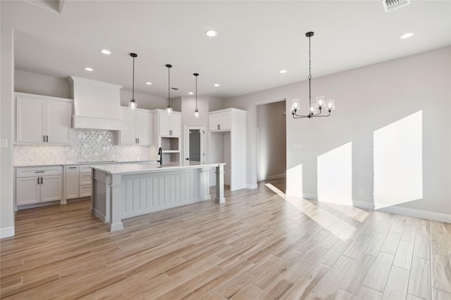 kitchen featuring custom exhaust hood, an inviting chandelier, white cabinets, a center island with sink, and decorative light fixtures