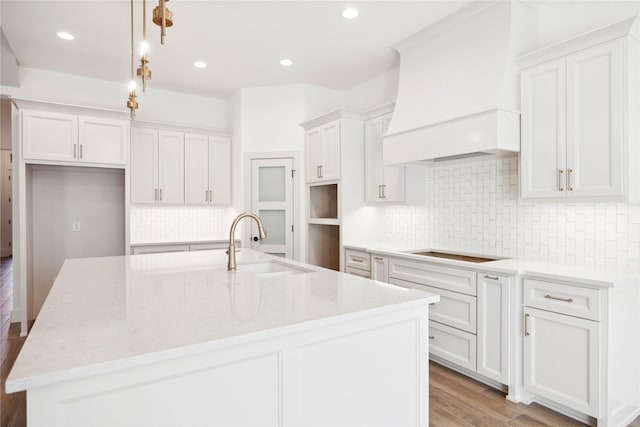 kitchen with premium range hood, hanging light fixtures, an island with sink, cooktop, and white cabinetry