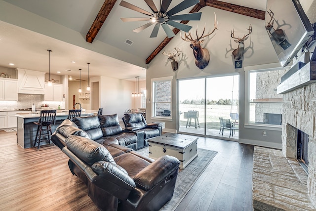 living room featuring ceiling fan, beam ceiling, high vaulted ceiling, light hardwood / wood-style floors, and a stone fireplace