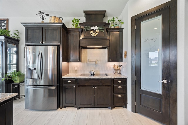 kitchen with stainless steel refrigerator with ice dispenser, backsplash, black electric stovetop, light stone counters, and dark brown cabinetry