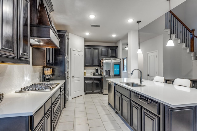 kitchen featuring premium range hood, sink, decorative light fixtures, appliances with stainless steel finishes, and a kitchen island with sink