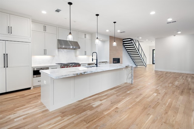 kitchen with a kitchen island with sink, ventilation hood, white cabinets, hanging light fixtures, and light stone countertops