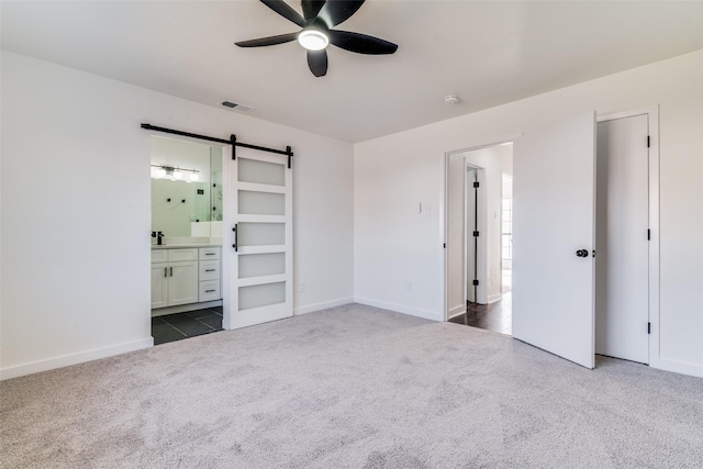 unfurnished bedroom featuring connected bathroom, a barn door, ceiling fan, and dark carpet