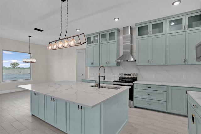 kitchen with stainless steel range with electric stovetop, wall chimney exhaust hood, a center island with sink, and light stone countertops