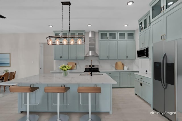 kitchen with light stone countertops, stainless steel fridge, wall chimney exhaust hood, hanging light fixtures, and an island with sink