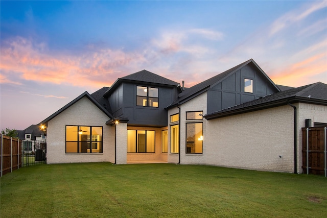 back of house at dusk with brick siding, a shingled roof, a lawn, central AC, and a fenced backyard