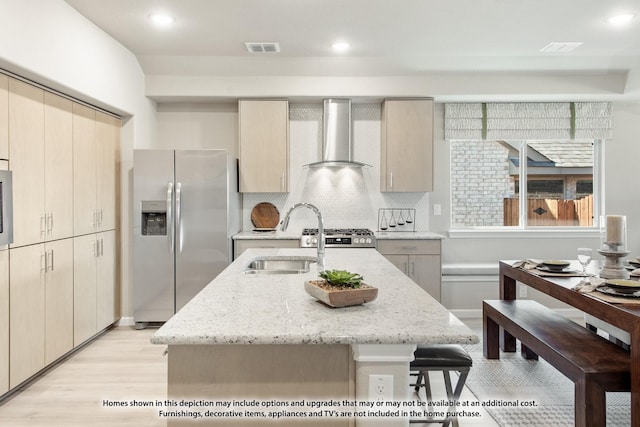 kitchen with light stone countertops, sink, wall chimney range hood, stainless steel fridge with ice dispenser, and an island with sink