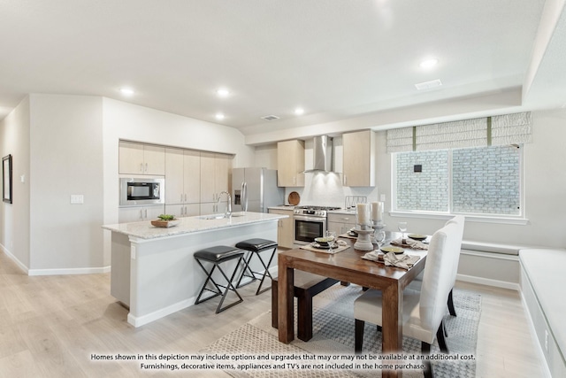 kitchen featuring light stone countertops, stainless steel appliances, wall chimney range hood, a kitchen island with sink, and light wood-type flooring