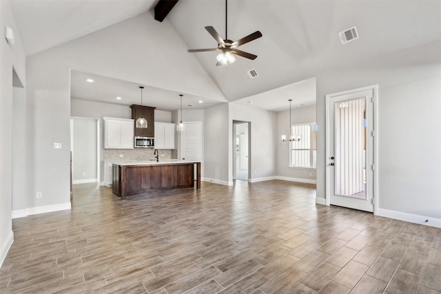 unfurnished living room with ceiling fan with notable chandelier, high vaulted ceiling, sink, beam ceiling, and light hardwood / wood-style flooring