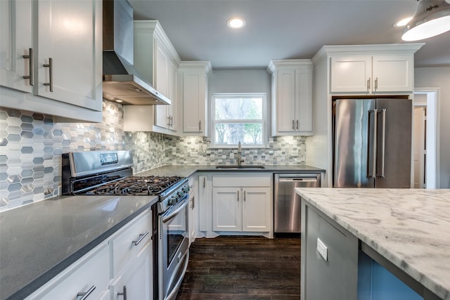 kitchen with appliances with stainless steel finishes, white cabinets, a sink, wall chimney range hood, and dark stone counters